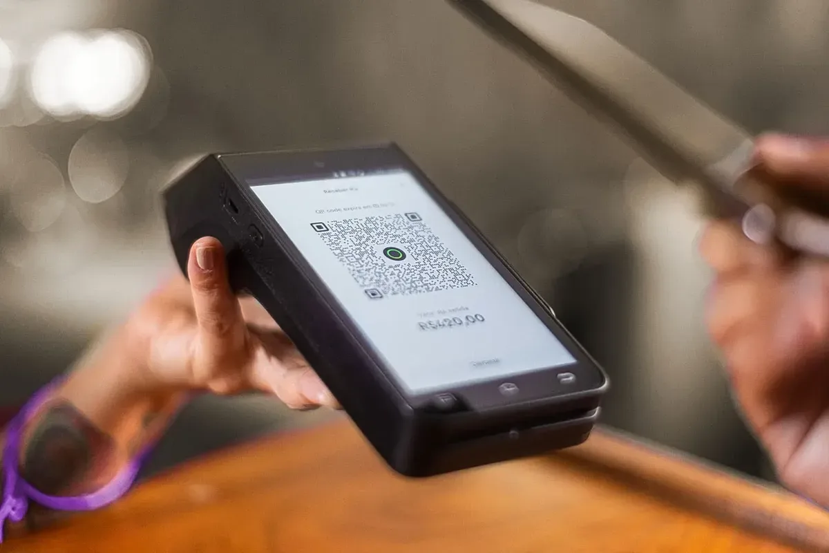 Pessoa segurando maquininha de cartão exibindo QR Code para pagamento via Pix no valor de R$ 420,00.