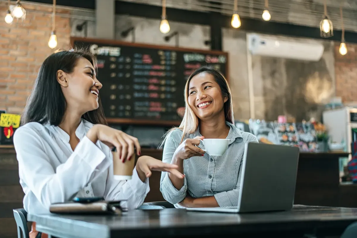 Duas mulheres sorridentes conversam em uma cafeteria aconchegante, tomando café e utilizando um laptop, representando networking e empreendedorismo em franquias.