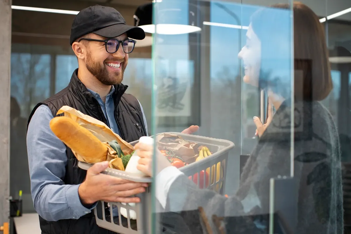 Entregador sorridente segurando uma cesta de compras com pães, leite, frutas e legumes, interagindo com uma cliente por meio de um balcão de vidro.